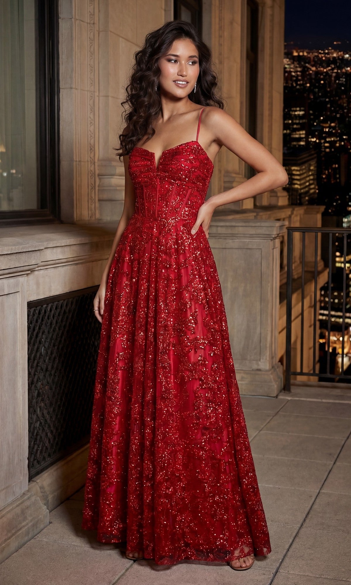 A woman stands on a balcony at night in a red Long Glitter Prom Dress: Blondie Nites 5409BN with thin straps and a fitted bodice. City lights twinkle behind her, highlighting her elegant formal evening gown.